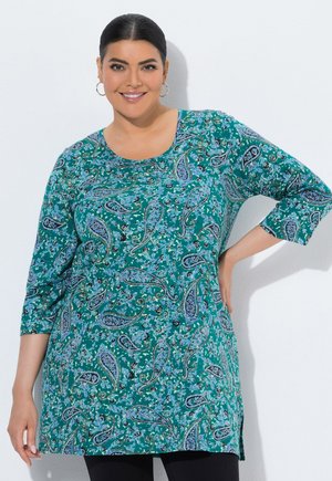 Smilende kvinde iført teal paisley-print tunika med trekvartærmer, stående med den ene hånd på hoften mod hvid baggrund.