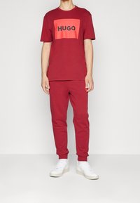 Camiseta de algodón roja con un logo cuadrado en naranja que presenta la palabra "HUGO". Combinada con pantalones de chándal rojos a juego y zapatillas blancas.