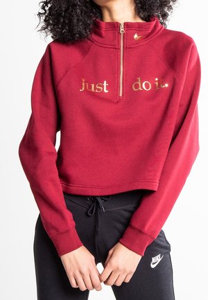 Woman wearing a cropped red zip-up sweatshirt reading "Just do it." and black Nike sweatpants with white logo, standing with hands clasped.