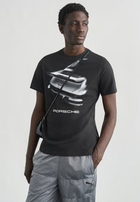 Μαύρο βαμβακερό t-shirt με ασημένιο γραφικό αυτοκινήτου και κείμενο "PORSCHE". Το μοντέλο φοράει γκρι ημι-επενδυμένα σορτς και μικρή μαύρη τσάντα.