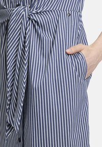 Abito di tessuto a righe blu e bianche con dettaglio in vita annodato, dotato di tasca laterale e texture liscia. Accentatura con bottone a pressione visibile.