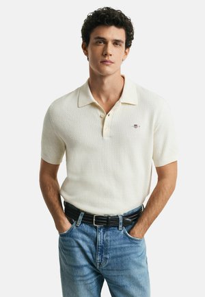 Korte mouw polo, witte textuurstof met een lichtbeige kraag, met een klein logo op de borst. Draagt met blauwe denim jeans.