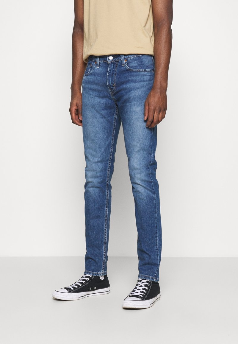 Personne portant un jean slim bleu, un t-shirt beige et des baskets montantes noires, debout devant un fond blanc uni.