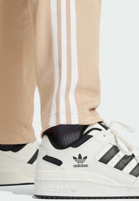 Beige athletic pants with white stripes and black accents are paired with white sneakers featuring black stripes and logo. Textured fabric visible.