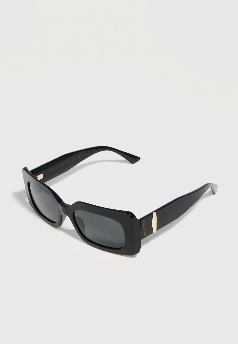 Μαύρα ορθογώνια γυαλιά ηλίου με σκούρους φακούς και χρυσές λεπτομέρειες στους παχείς βραχίονες, απεικονιζόμενα σε ανοιχτό φόντο.