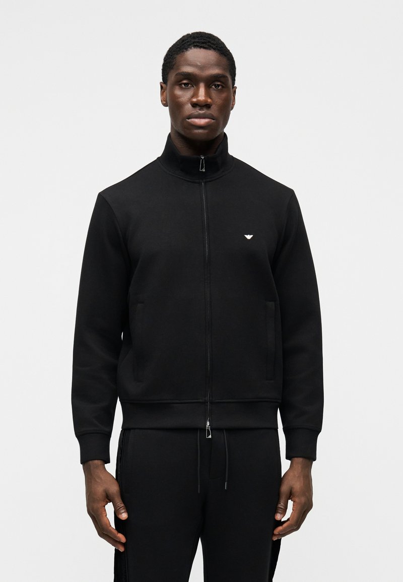 Juodas su užtrauktuku megztinis su aukštu apykakle, kišenėmis priekyje, ribinėmis rankogaliais ir logotipo detalėmis ant krūtinės. Minkštas audinys su lygiu paviršiumi.