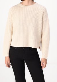 Suéter beige de punto con cuello redondo, textura acanalada y aberturas laterales, combinado con vaqueros negros. El suéter tiene mangas ligeramente oversize.