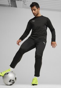 Zwarte sportieve trainingspak met lange mouwen en broek, met een Puma-logo. Neon gele voetbalschoenen en een zilveren voetbal zijn aanwezig.