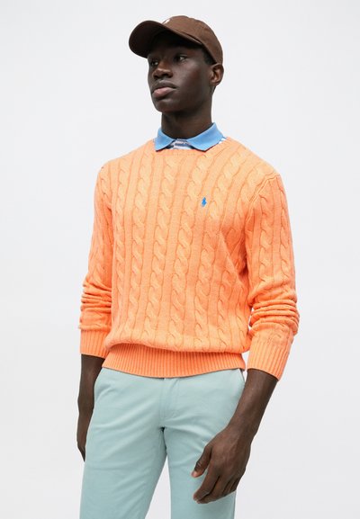 Πλεκτό πουλόβερ με πλεξούδα σε έντονο πορτοκαλί, με ριμπ μανσέτες και τελείωμα. Συνδυασμένο με ανοιχτό μπλε πουκάμισο με γιακά και παντελόνι μέντας. Καφέ καπέλο.