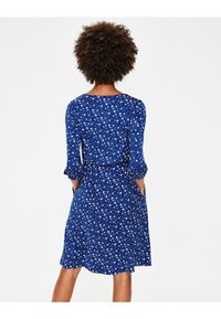 Blå blommig klänning med trekvartsärmar och fickor, med en figursydd midja och utflugen kjol, tillverkad av mjukt tyg med en texturerad yta.