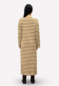 Lange gestreepte jurk met bruine en crèmekleurige strepen, gemaakt van zachte stof. Heeft een ronde halslijn en lange mouwen. Gecombineerd met zwarte laarzen.