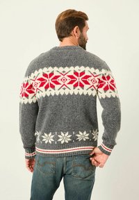 Mann in einem grauen Strickpullover mit weißen und roten Schneeflockenmuster, kombiniert mit blauen Jeans, steht mit dem Rücken zur Kamera.