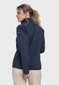 Marineblaue Softshell-Jacke mit hohem Kragen, Reißverschlusstaschen und tailliertem Design. Glatte Textur mit Schöffel-Logo auf dem Ärmel.