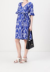 Blå blommönstrad klänning med korta volangärmar, insnörd midja, parat med vita klacksandaler med blockklack och en svart handväska.