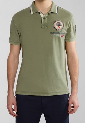 Mand iført olivengrøn poloshirt med krave og lapper, kombineret med mørkeblå bukser mod en ensfarvet baggrund.