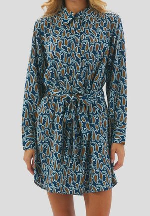 Femme portant une robe boutonnée à manches longues avec un motif cachemire bleu, marron et blanc et une ceinture nouée à la taille.
