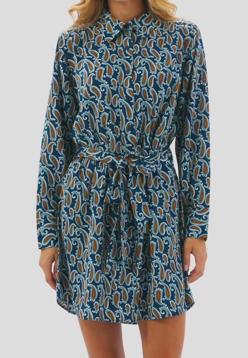Femme portant une robe boutonnée à manches longues avec un motif cachemire bleu, marron et blanc et une ceinture nouée à la taille.