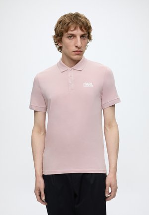 Homme aux cheveux bouclés portant un polo rose clair Karl Lagerfeld et un pantalon noir, debout devant un fond blanc uni.