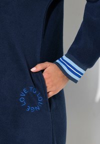 Pigiama in pile navy con tasca laterale. Il polsino presenta strisce orizzontali di blu chiaro e blu scuro. Il testo ricamato dice "ADORE LOUNGE."