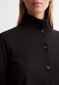 Schwarzes Langarm-Oberteil mit hohem Kragen aus weichem Stoff, mit einer Knopfleiste vorne und sichtbaren schwarzen Knöpfen, glatte Textur mit gerippten Akzenten.