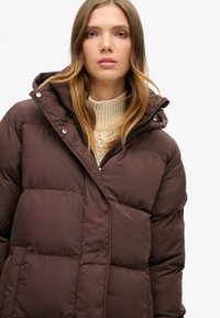 Brun pufferjakke med quiltet design, der har hætte, trykknapper og lynlås foran. Varm, letvægts materiale.