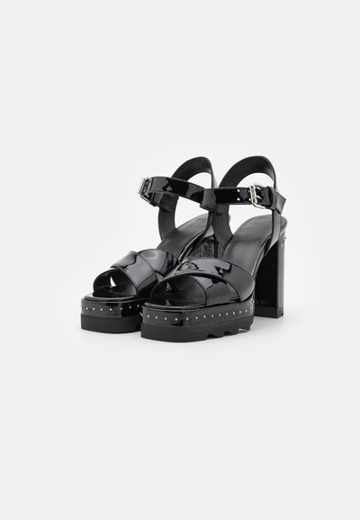 Sandalias de plataforma de charol negro con tacón grueso, múltiples correas y detalles de tachuelas plateadas en la suela. Hebilla ajustable en el tobillo.