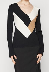 Sort strikket top med V-hals, der har geometriske farveblokke i creme, tan og sort. Lange ærmer og tætsiddende design. Glat tekstur.