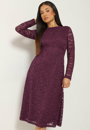 Femme aux longs cheveux lisses portant une robe midi en dentelle violette ajustée à manches longues, posant avec une main derrière la tête.