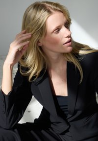 Szőke nő fekete blézerben, oldalra nézve, kezét a hajához emelve, természetes fény világítja meg, egyszínű háttér előtt.