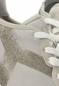 Zapatilla gris con acentos plateados brillantes en el lateral, cordones blancos y un collar acolchado. Parte superior texturizada con acabados suaves y de ante.