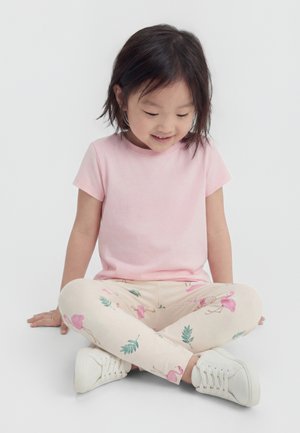 Młoda dziewczyna siedzi ze skrzyżowanymi nogami, ubrana w jasnoróżową koszulkę, kremowe legginsy z nadrukiem różowych flamingów i zielonych liści oraz białe trampki.