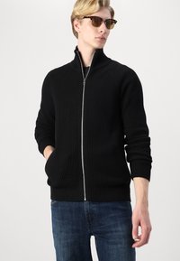 Svart ribbestrikket genser med høy krage, sølvglidelås og sidelommer, kombinert med blå jeans og skilpaddemønstrede solbriller.
