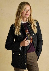 Jachetă neagră matlasată cu accente aurii, purtată peste un pulover burdund cu un design de vulpe, asortată cu pantaloni în carouri.