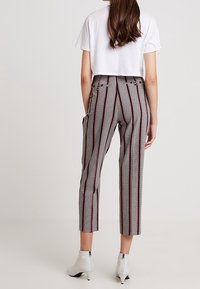 Kvinna som bär mönstrade, korta byxor med vertikala ränder och vita ankelstövlar, stående mot en enfärgad bakgrund, sett bakifrån.