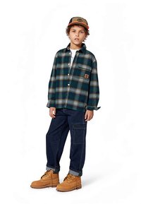 Cămașă carourată verde și bleumarin cu buzunar la piept, pantaloni din denim închis la culoare și cizme căptușite maro. Accesorizată cu o șapcă de baseball cu un logo.