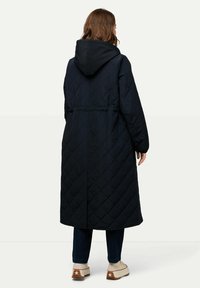 Lång, marinblå quiltad kappa med huva, elastisk midja och sidofickor. Tyget har ett diamantmönster för extra textur.