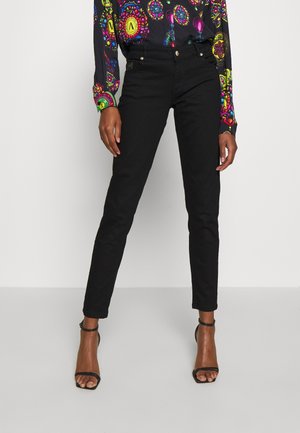 Kvinde iført sorte slim-fit bukser, farverig mønstret langærmet skjorte og sorte åbne tå-sandaler med ankrem, stående mod hvid baggrund.