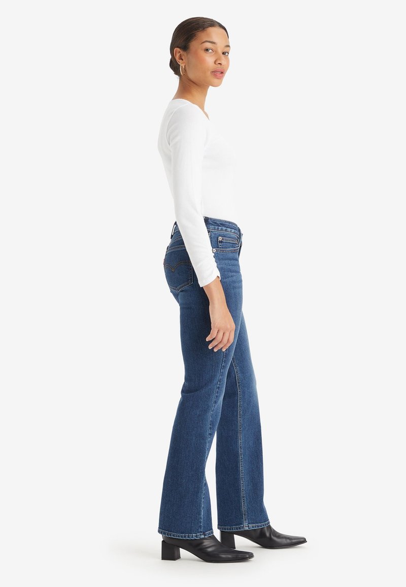 Donna che indossa un top a maniche lunghe bianco, jeans a gamba bootcut blu e stivali con tacco neri, in piedi di lato su uno sfondo bianco.