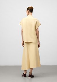 Donna con i capelli raccolti in un bun, che indossa un top beige a maniche corte e una gonna lunga abbinata, con le spalle rivolte verso di noi, abbinata a sneakers marrone scuro.
