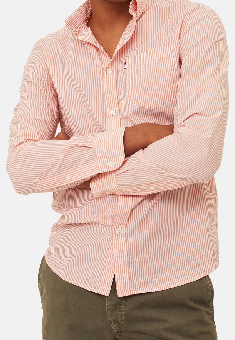 Homme portant une chemise boutonnée rayée rose et blanche avec poche poitrine, bras croisés, et pantalon vert olive sur fond uni.