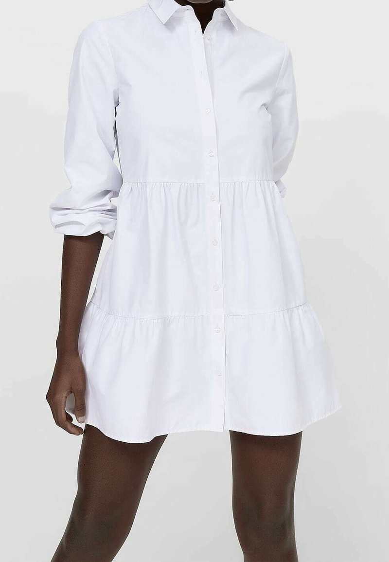 Femme portant une robe mini blanche boutonnée à manches longues retroussées jusqu'aux coudes, sur un fond uni.