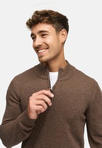 Brun zip-up tröja med ribbad krage och muddar, med en mjuk texturerad stickning. Modellen håller i dragfliken och ler.