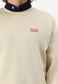 Beige színű pulóver bordázott kerek nyakú kivitelben. A bal felső mellkas részen egy kiemelkedő piros és fekete "HUNTER" logó található.