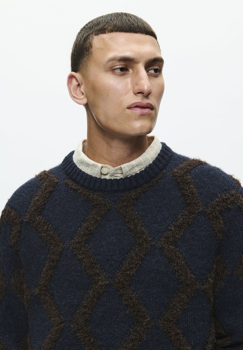 Pull bleu marine avec des motifs losanges marron, col rond et tricot texturé. Porté par-dessus une chemise claire à col.