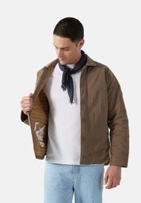 Giacca marrone trapuntata con chiusura a bottone, dotata di un esterno testurizzato e di un rivestimento interno con un design stampato. Indossata sopra una camicia bianca.