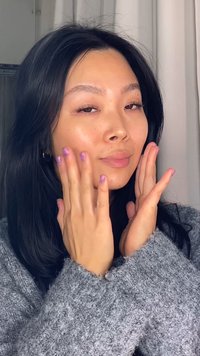 Junge Frau mit langen schwarzen Haaren, die einen grauen Pullover trägt und sanft ihre Wangen berührt, dabei eine glatte Haut und glitzernden lila Nagellack zeigt.