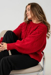 Sarkans adīts džemperis ar kabeli, ribotām piedurknēm un brīvu siluetu, kombinēts ar melnajām biksēm. Gluda faktūra, masīvs izskats.