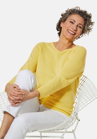 Gelber Strickpullover mit langen Ärmeln, Rundhalsausschnitt und gerippten Bündchen, kombiniert mit weißen, figurbetonten Hosen, vor einem schlichten Hintergrund.