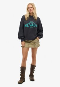 Sudadera gris de gran tamaño con texto en color azul verdoso que dice "Sierra Nevada". Combinada con una falda cargo color beige y botas marrones hasta la rodilla con hebillas.