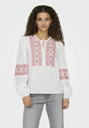 Blouse blanche avec des motifs rouges brodés sur le devant et les manches, dotée d'une fermeture à nouer et de manches longues et volumineuses.